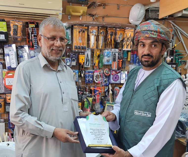 Two men standing in the shop, holding a Consumer Protection Authority plaque in its presentation box between them.